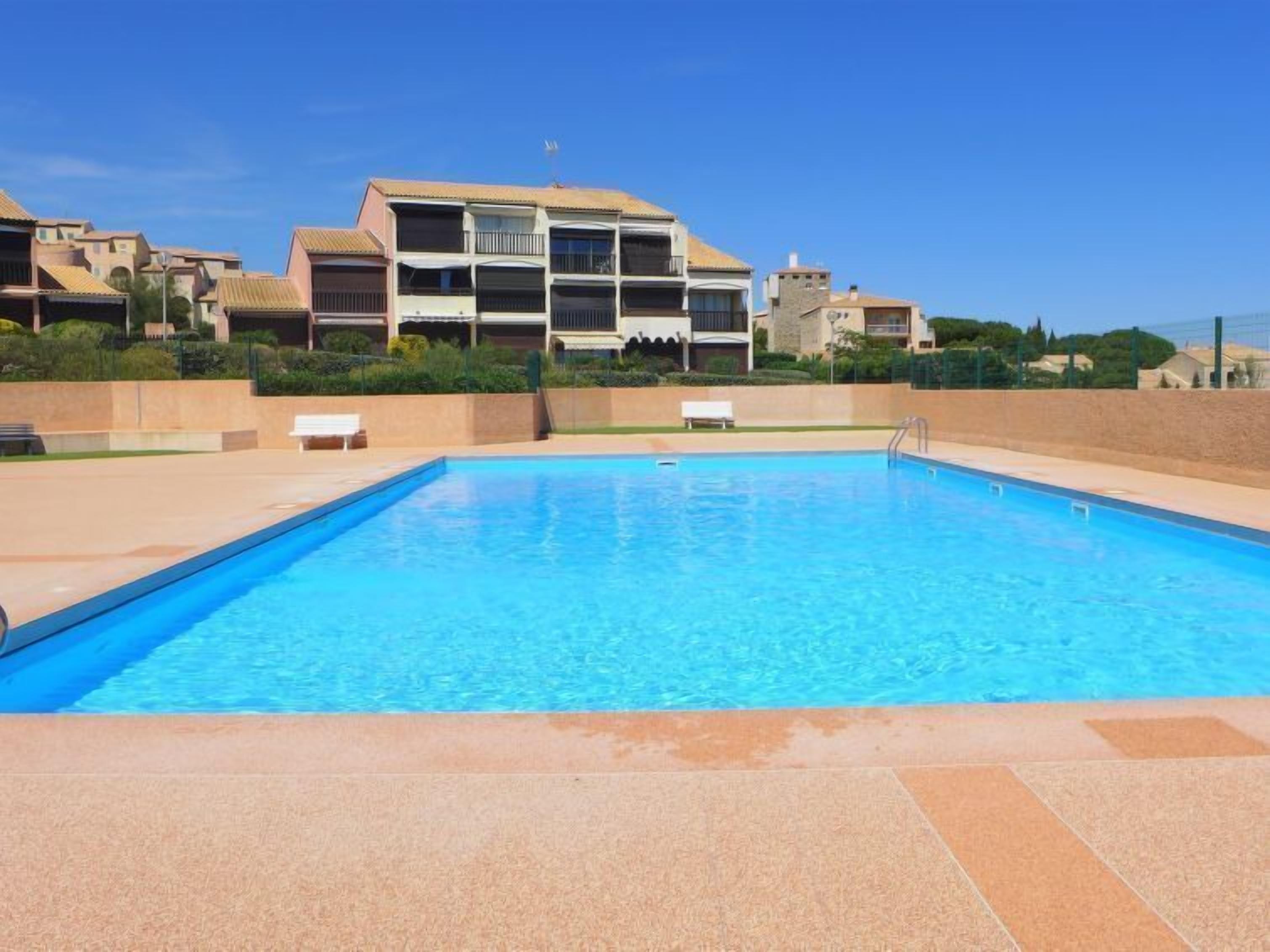 Une piscine rectangulaire à l’eau d’un bleu limpide, entourée d’une terrasse carrelée. Au fond, on aperçoit des immeubles à plusieurs étages dotés de balcons, et un ciel d’un bleu éclatant.