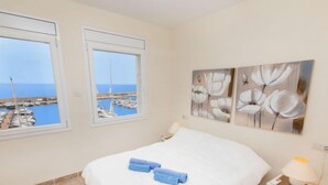 Un dormitorio con una cama, dos ventanas con vista al puerto deportivo y tres cuadros florales en la pared.