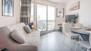 A modern living room with a sofa, a dining table with chairs, a TV, and a balcony with a view. - Copacabana by Interhome (Canet-Plage)