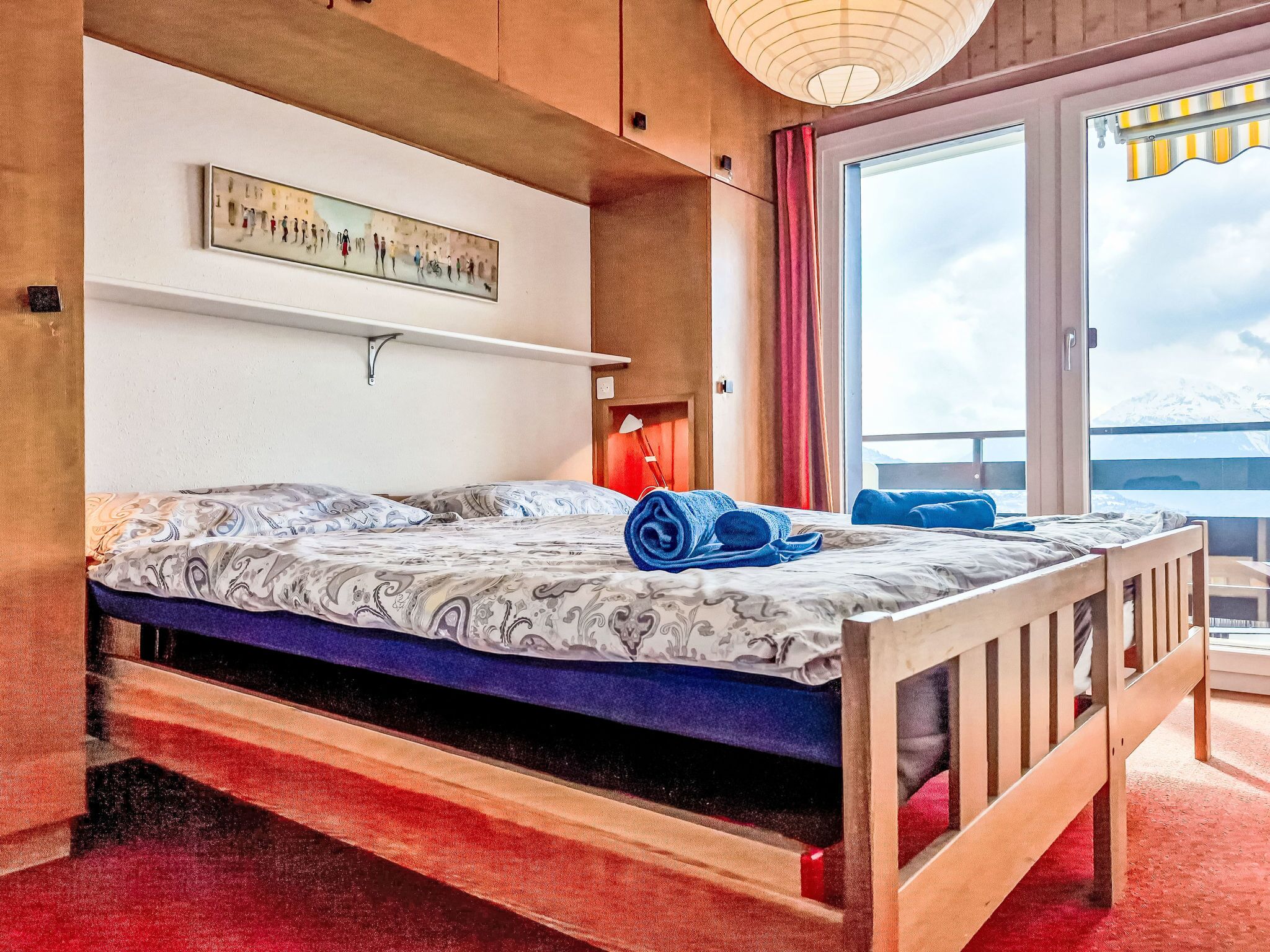 Une chambre à coucher avec un lit, une étagère fixée au mur, une fenêtre avec des rideaux et une vue sur l’extérieur.