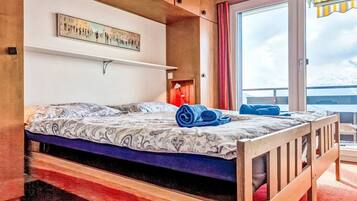 Une chambre à coucher avec un lit, une étagère fixée au mur, une fenêtre avec des rideaux et une vue sur l’extérieur.