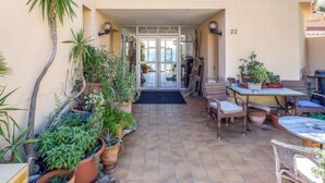 Une cour avec un sol carrelé, des plantes en pot, du mobilier d’extérieur et une porte donnant sur un espace intérieur.