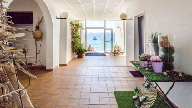 Un couloir au sol carrelé mène à une porte vitrée qui donne sur un balcon offrant une vue sur la mer.