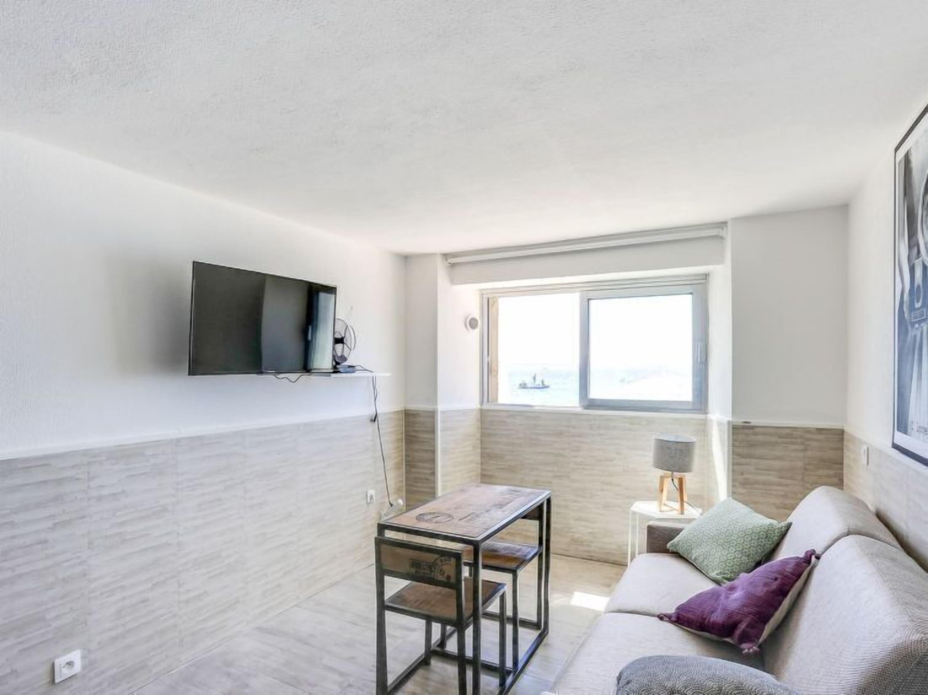 Un salon moderne avec un téléviseur à écran plat fixé au mur, une petite table en bois, un canapé avec des coussins et une fenêtre donnant sur la mer.