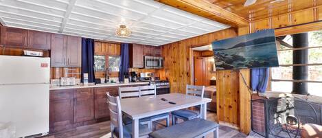 Une cuisine de style rustique avec des meubles en bois, une table à manger avec des bancs, une grande fenêtre donnant sur l’extérieur et une cheminée avec un paravent.