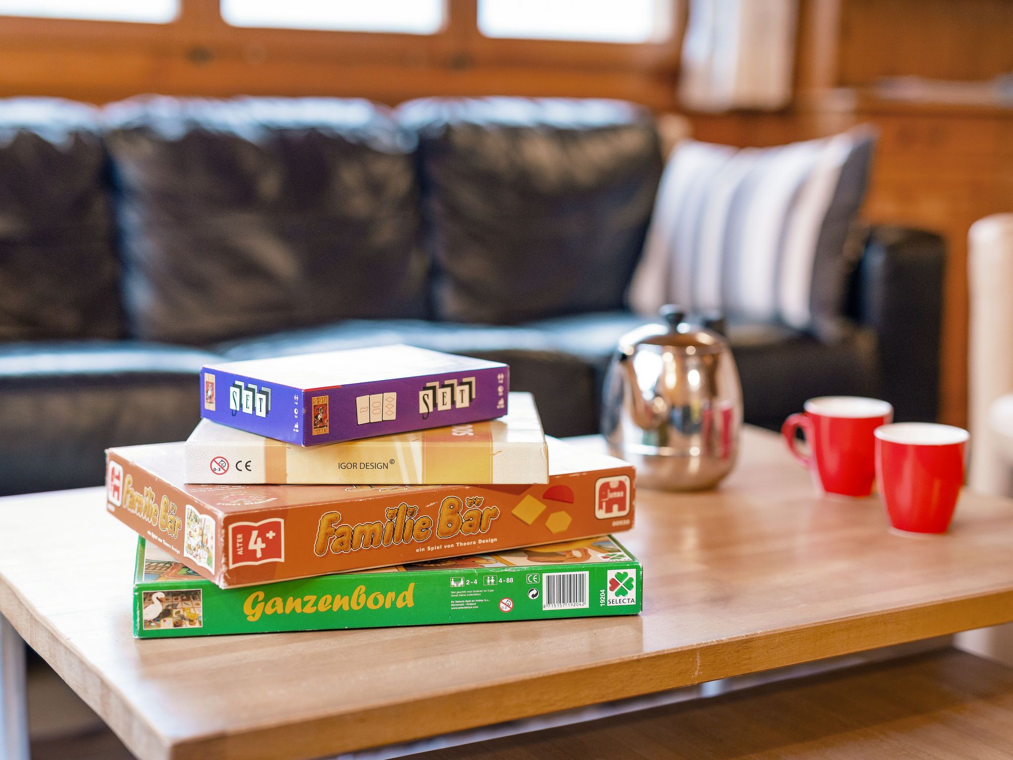 Un montón de juegos de mesa sobre una mesa de madera, con una cafetera y dos tazas rojas al fondo.