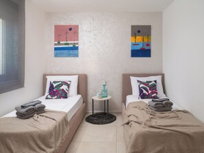 Habitación doble con dos camas, cada una con almohada y manta, una mesita de noche con un florero, y una obra de arte en la pared.
