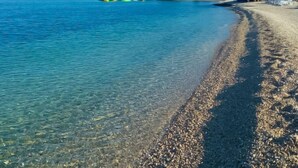 Ein Strand mit klarem Wasser, Kieselsteinen und ein Gebäude im Hintergrund, auf dem 'Beach Bar' steht.