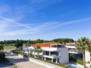 En moderne boligbygning med swimmingpool, omgivet af grøn vegetation og en klar himmel.