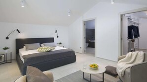 Une chambre moderne avec un grand lit, des tables de chevet, un bureau, une chaise, une petite table ronde avec des fruits et un dressing.