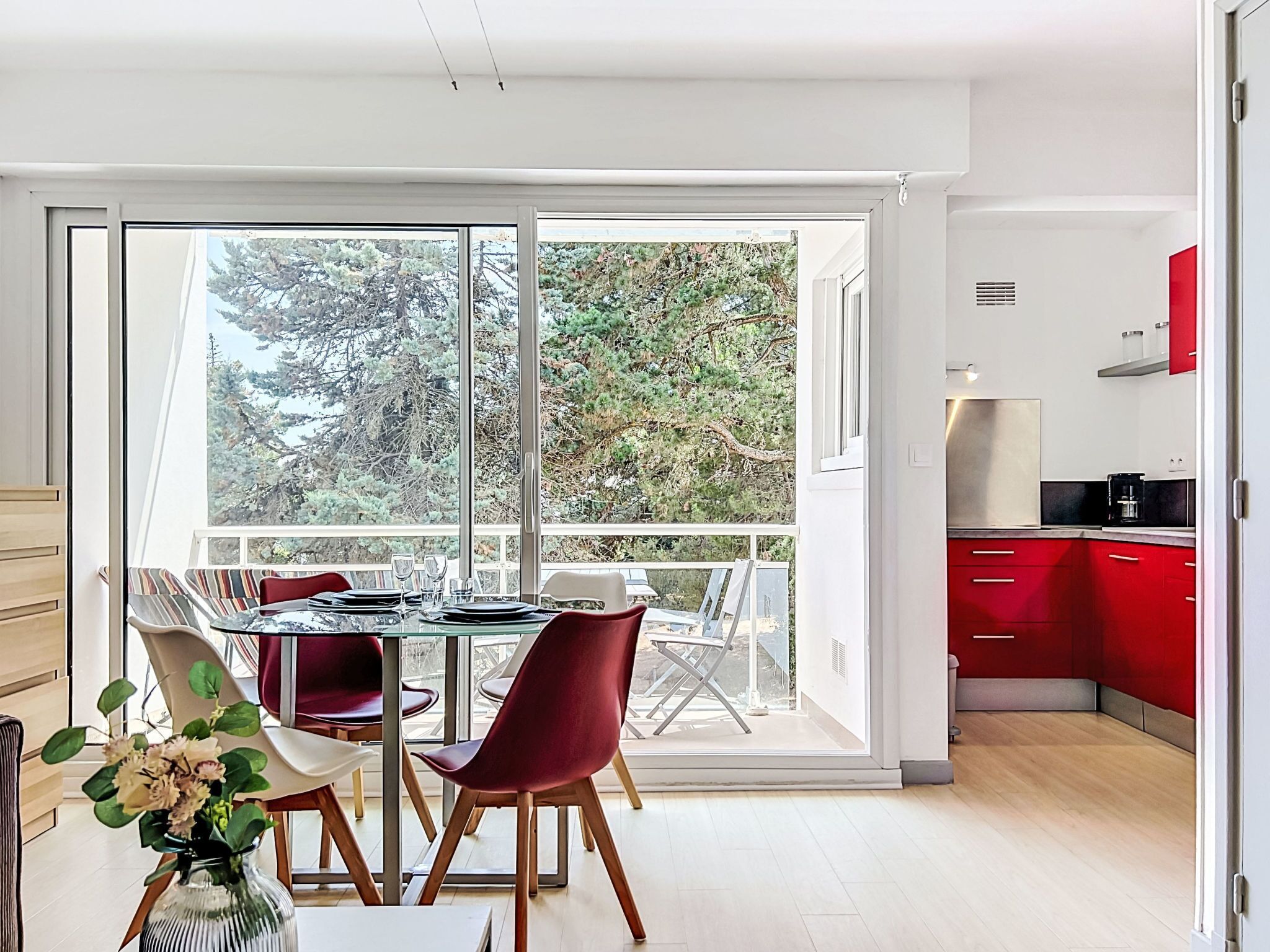 Una cocina moderna con armarios rojos, un comedor con mesa y sillas de vidrio, y vistas a árboles a través de amplias ventanas.