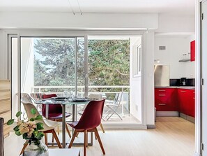 A modern kitchen with red cabinets, a dining area with a glass table and chairs, and a view of trees through large windows. - Port An Dro by Interhome (Carnac)