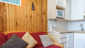 Een compacte kamer met een houten wand, een rode bank met kussens, een kleine keuken met een magnetron en een koffiezetapparaat, en een witte vaatwasser.