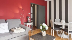A modern living room with a grey sofa, a small table with a vase of flowers, a dining area with a white table and chairs, and a red accent wall. - Junior Suite by Interhome (Ascona)