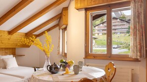 A cozy room with a wooden ceiling, a round table with a floral tablecloth, a vase with yellow flowers, and a window with a view of a building and greenery outside. - Eldorado 321 by Interhome (Verbier)