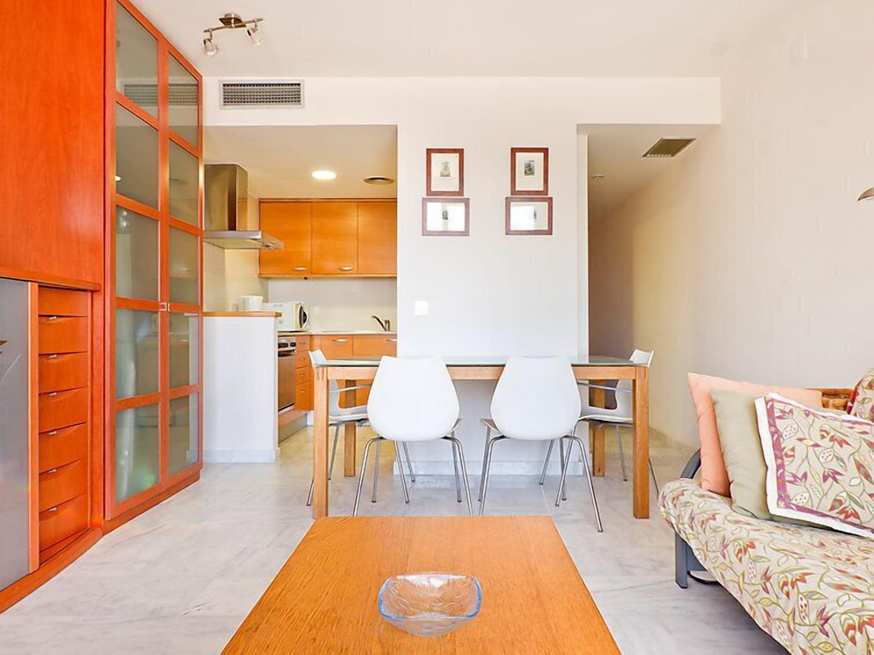 A compact living space with a kitchenette, a dining area with two chairs, a sofa with patterned cushions, and a glass coffee table.