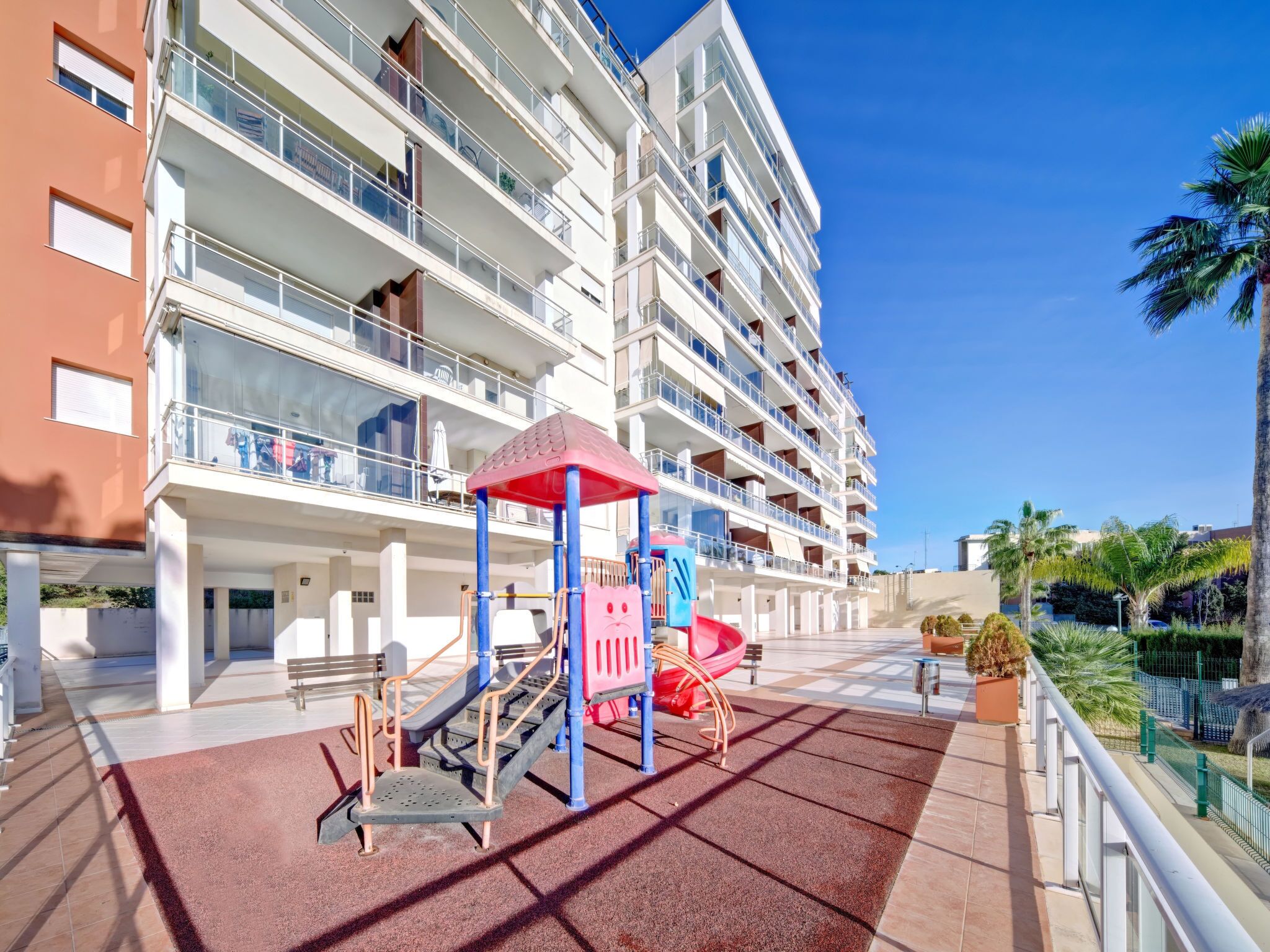Un immeuble résidentiel à plusieurs étages doté de balcons, d’une aire de jeux avec des toboggans et des structures d’escalade, et un ciel bleu dégagé.
