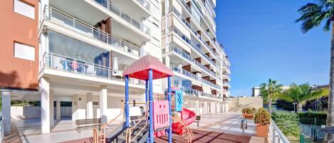 Un immeuble résidentiel à plusieurs étages doté de balcons, d’une aire de jeux avec des toboggans et des structures d’escalade, et un ciel bleu dégagé.
