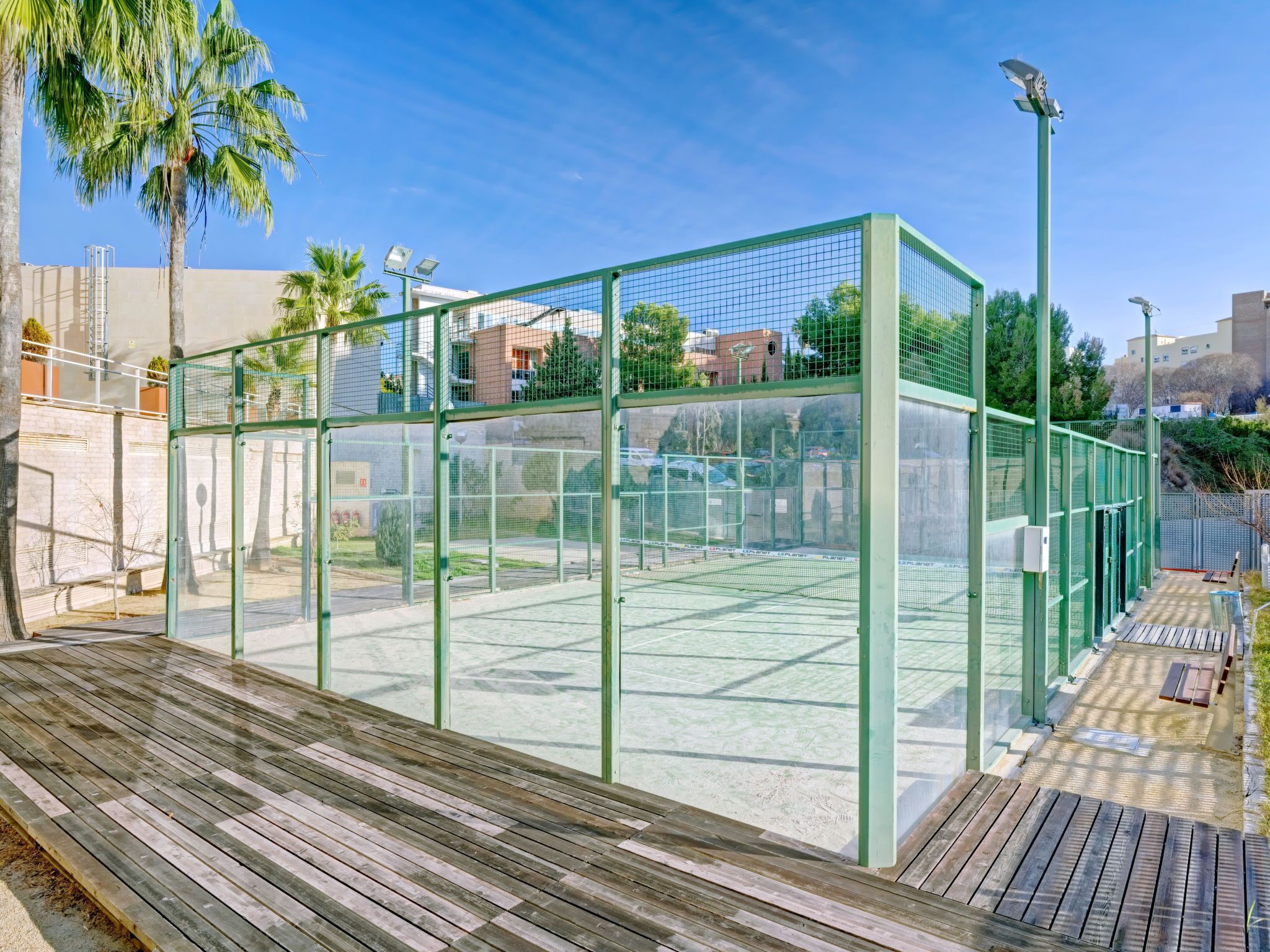 A tennis court enclosed by a green metal fence with a wooden deck in front.