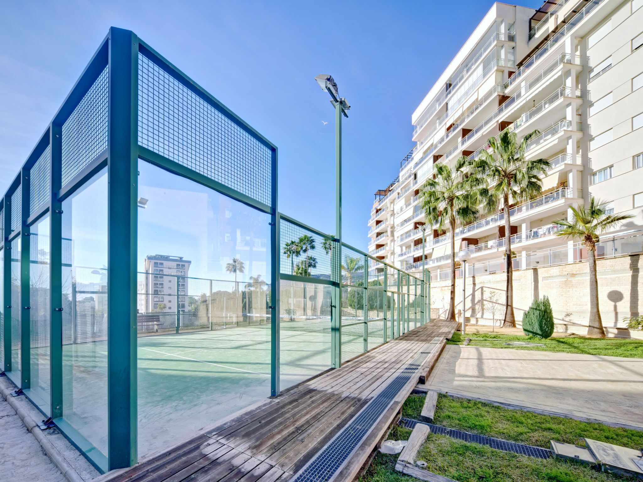 A tennis court with a glass enclosure, adjacent to a multi-story residential building with balconies and palm trees.