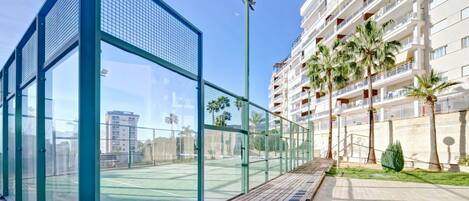 A tennis court with a glass enclosure, adjacent to a multi-story residential building with balconies and palm trees.