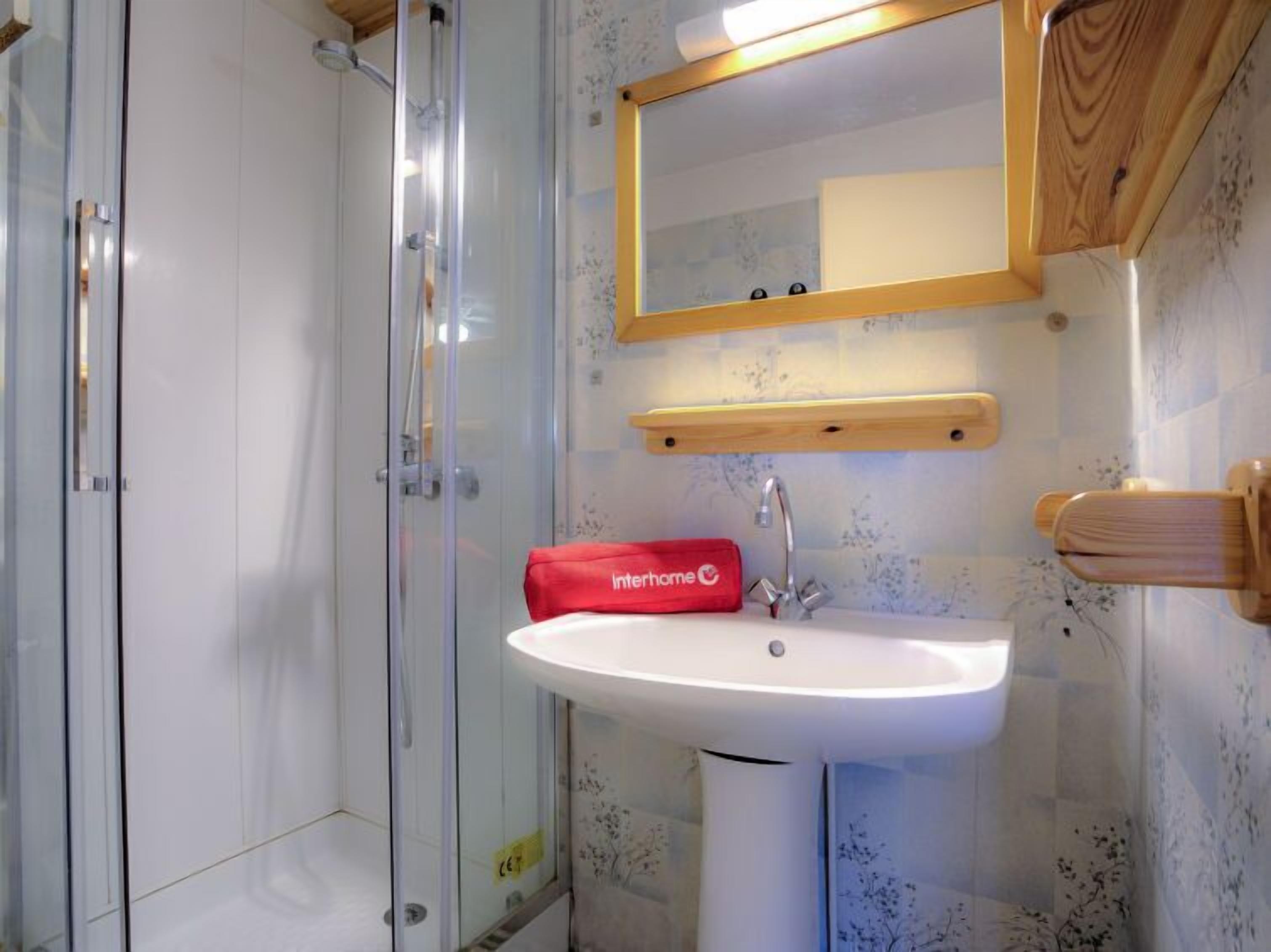 A bathroom with a glass shower enclosure, a white sink, a mirror with a wooden frame, and a red Interhome towel.