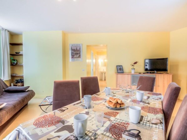 Un coin repas avec une table dressée pour quatre personnes, un téléviseur posé sur un meuble et une bibliothèque contenant divers objets.
