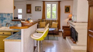 A kitchen with a bar stool, a dining area with a sofa, a fireplace, and a window with curtains. - Les Hauts de Palayson by Interhome (Le Muy)
