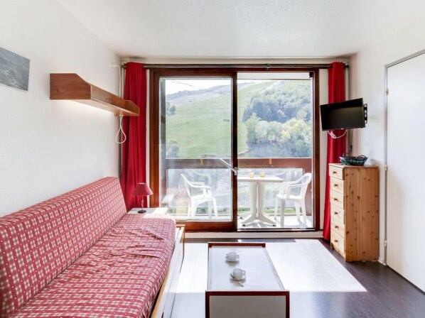 A room with a red patterned sofa, a small table with cups, a TV, and a view of a green landscape through a sliding door.