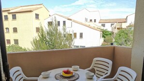 Un balcon avec une table ronde et quatre chaises blanches, un saladier rempli de fruits et un set de table en fibres tressées.