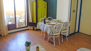 Un coin repas avec une table dressée pour quatre personnes, entouré de chaises blanches ornées de coussins à motifs. Un grand miroir reflète la pièce, et un buffet jaune est visible au fond.
