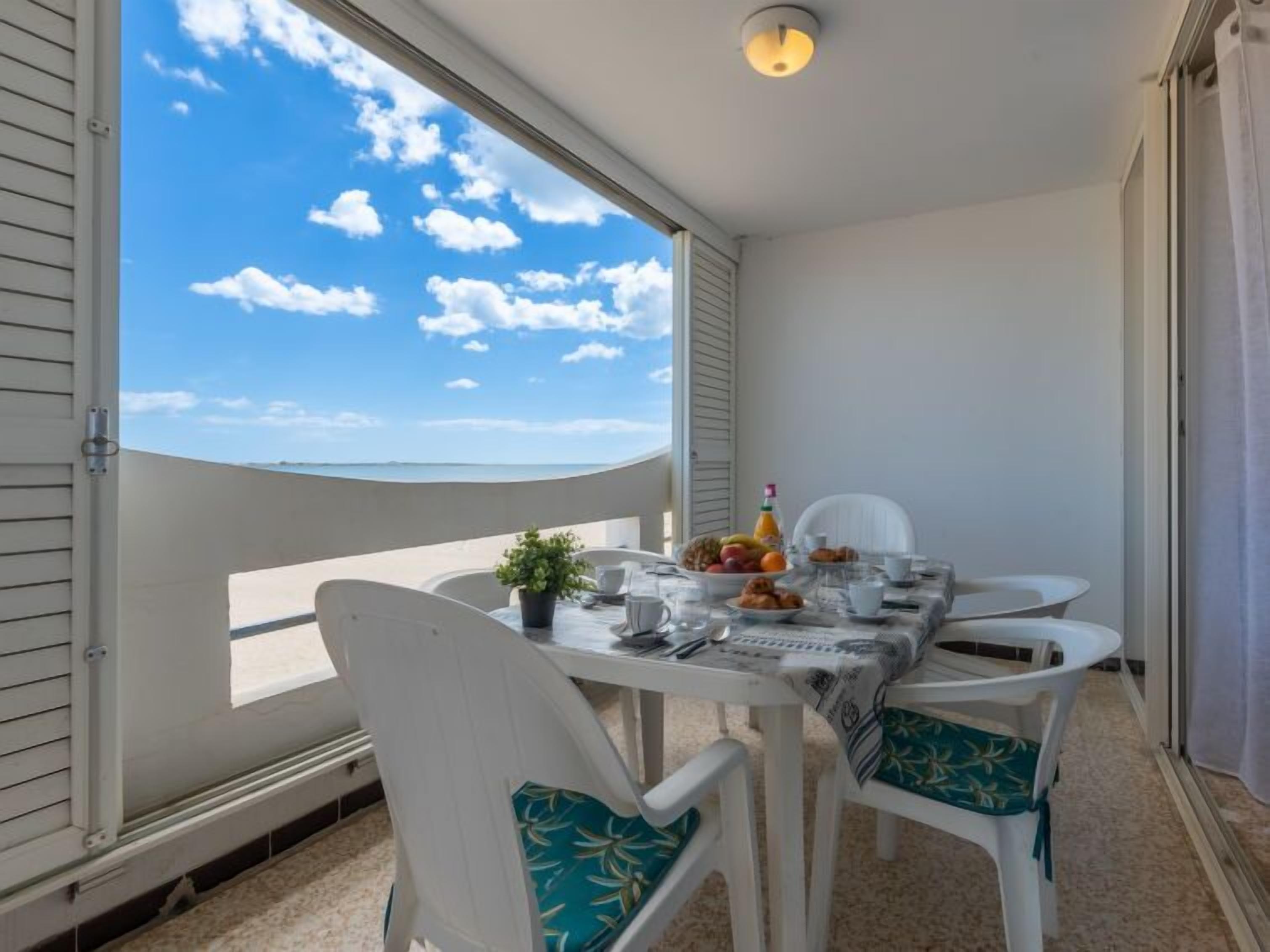 A balcony with a table set for breakfast, white chairs with teal cushions, and a clear view of the sea.