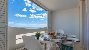 Un balcon avec une table dressée pour le petit-déjeuner, des chaises blanches ornées de coussins turquoise et une vue dégagée sur la mer.