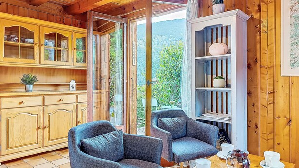 A cozy cabin interior with wooden walls and ceiling, a dining area with two blue armchairs and a table set with cups and saucers, and a kitchen with wooden cabinets and a potted plant.