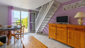 Un espace repas comprenant une table et des chaises, un banc en bois, un téléviseur posé sur un meuble en bois, et un escalier menant à un espace extérieur.