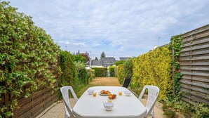 Une terrasse avec une table et des chaises blanches, entourée d’une clôture en bois et de verdure.