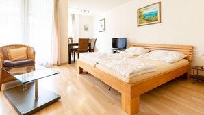 Habitación de hotel con una cama de madera, una mesa de centro de vidrio, una silla de mimbre, un escritorio con silla, un televisor y un cuadro en la pared.