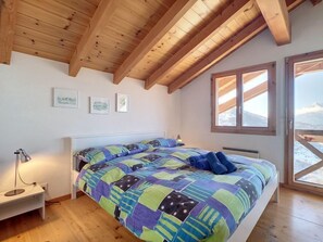 A bedroom with a wooden ceiling, a bed with a colorful quilt, a bedside table with a lamp, and a window with a mountain view.