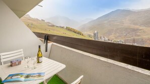 Varanda com mesa posta para dois, uma garrafa de vinho e um folheto. A vista inclui uma paisagem montanhosa e o horizonte de uma cidade ao longe.