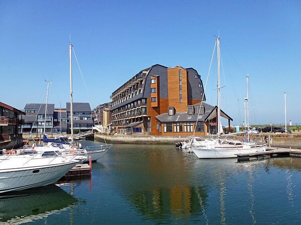 A marina with several boats docked, a modern building with multiple floors and balconies, and a clear blue sky.