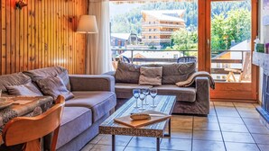 A cozy living room with a wooden wall, a sofa, a coffee table with glasses, and a view of a mountain village through a glass door. - Les Platanes E3 by Interhome (Nendaz)