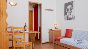 A small, cozy room with a wooden dining table and chairs, a bed with striped bedding, a small bedside table with a lamp, and a red curtain in the background. - Rond Point des Pistes (Val Claret) by Interhome (Tignes)