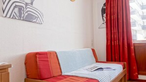 A room with a wooden sofa bed, red and white cushions, a striped blanket, and a wall-mounted light. - Rond Point des Pistes (Val Claret) by Interhome (Tignes)
