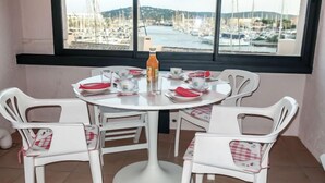 Een eethoek met een ronde tafel en vier witte stoelen, een fles sinaasappelsap, en uitzicht op een jachthaven met boten.