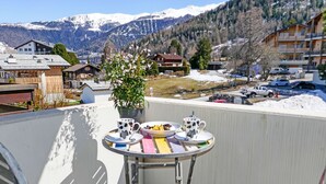 A balcony with a table set for two, a potted plant, and a view of a snowy mountain landscape. - Grand Place 5 by Interhome (Nendaz)