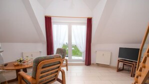 A spacious room with a dining table, a wooden chair, a television on a stand, and a view of greenery outside. - Meerblick by Interhome (Norddeich)