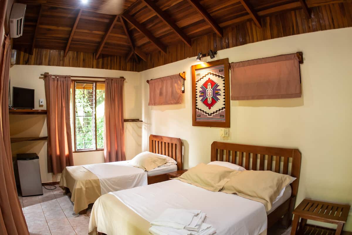 A room with two beds, a ceiling fan, a window with curtains, a wall-mounted TV, and a decorative wall hanging.