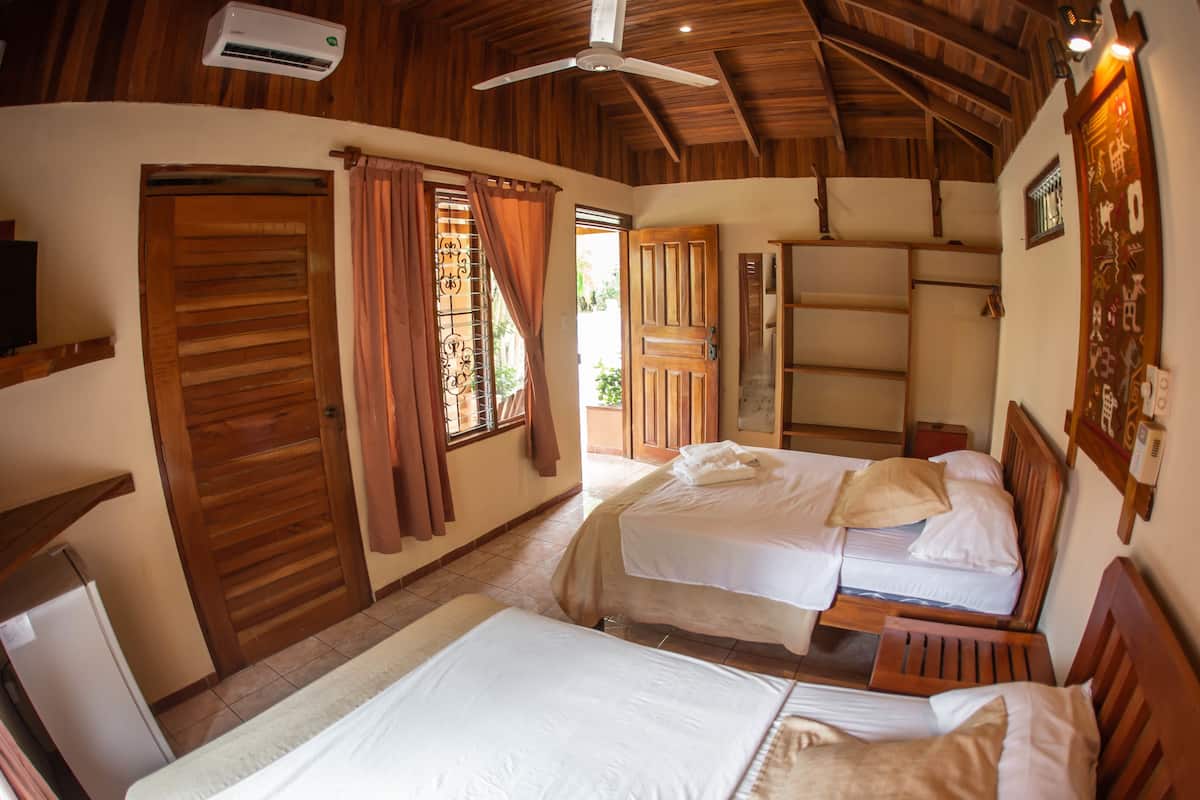 A room with two beds, wooden ceiling beams, a ceiling fan, an air conditioning unit, and a door with a window.