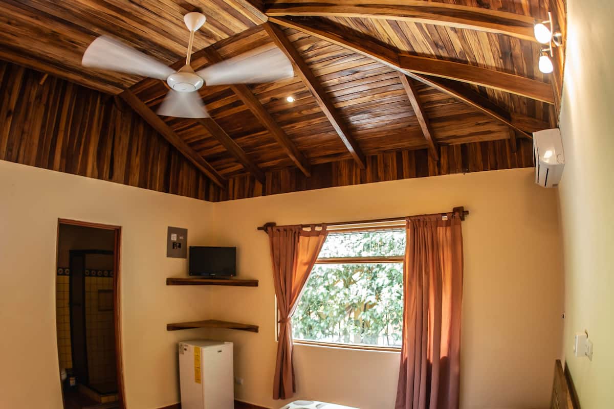 A bedroom with a wooden ceiling, a bed with white bedding, a small TV on a shelf, and a window with curtains.