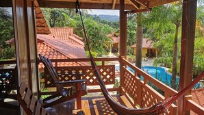 Un balcone in legno con un'amaca, una piscina e vista su un paesaggio tropicale.